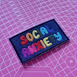 Può includere: Una toppa rettangolare in denim con la scritta ricamata "SOCIAL ANXIETY" in vari colori. La toppa ha un bordo blu scuro. È posizionata su un tappetino da taglio rosa con linee di griglia e misure.