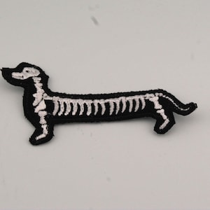 Può includere: Toppa ricamata in bianco e nero a forma di cane bassotto. Lo scheletro del cane è raffigurato con filo bianco su sfondo nero. La toppa è progettata per essere stirata o cucita sul tessuto.