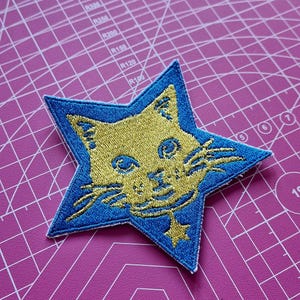 Peut inclure: Écusson brodé en forme d'étoile avec une bordure bleue et un motif de visage de chat doré. Le chat a des moustaches et un petit pendentif en forme d'étoile. L'écusson est sur un tapis de coupe rose.