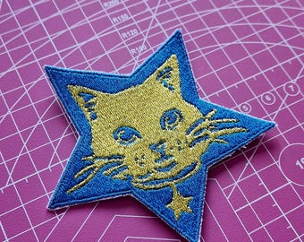 May include: Embroidered patch in the shape of a star with a blue border and a gold cat face design. The cat has whiskers and a small star pendant. The patch is on a pink cutting mat.