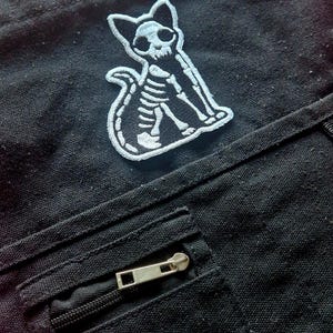 May include: Black canvas shoulder bag with a white embroidered cat skeleton patch. Features a zippered front pocket and a black shoulder strap. The bag has a tag that reads "FASHION INSIDER".