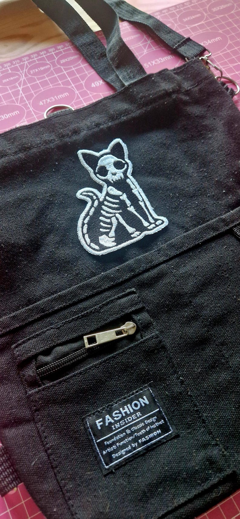 May include: Black canvas shoulder bag with a white embroidered cat skeleton patch. Features a zippered front pocket and a black shoulder strap. The bag has a tag that reads "FASHION INSIDER".