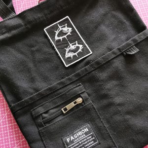 May include: Black canvas tote bag with a rectangular patch featuring two stylized white eyes. The bag has a front pocket with a zipper and a label that reads "FASHION INSIDER". The bag has a shoulder strap and a top handle.
