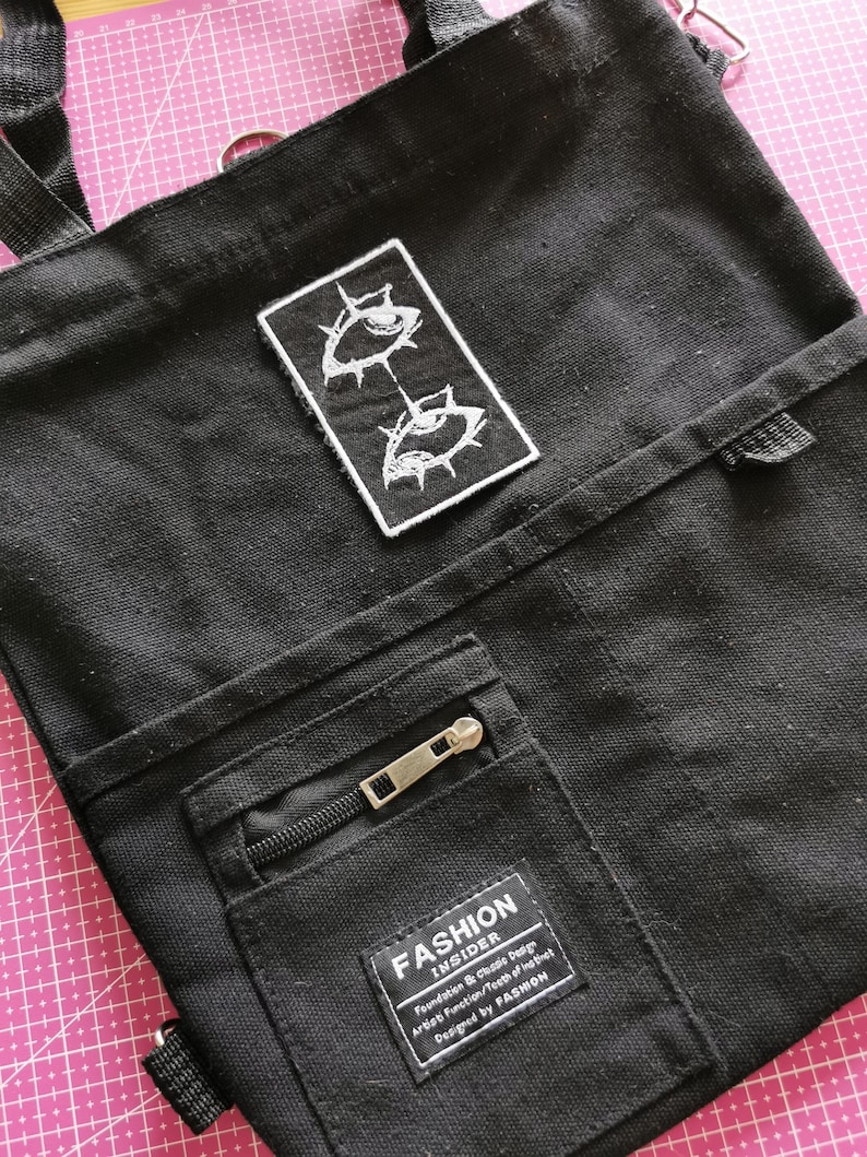 May include: Black canvas tote bag with a rectangular patch featuring two stylized white eyes. The bag has a front pocket with a zipper and a label that reads "FASHION INSIDER". The bag has a shoulder strap and a top handle.