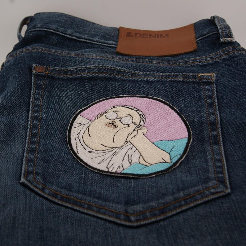 May include: Dark wash denim jeans with a patch on the back pocket. The patch features an embroidered illustration of a person with glasses, resting their head on their hand, within a pink and blue oval.