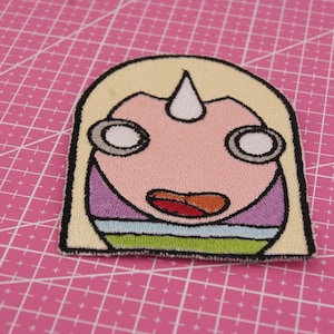 Könnte beinhalten: Gestickter Aufnäher mit einem Cartoon-Gesicht und einem Einhornhorn. Das Design umfasst einen hellgelben Hintergrund, ein rosa Gesicht und eine schwarze Umrandung. Die Augen sind weiß mit grauen Umrissen, und der Mund ist offen und zeigt eine rote Zunge.