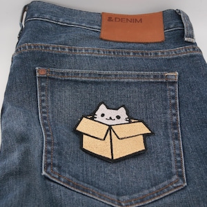Peut inclure: Jean bleu foncé avec un patch chat dans une boîte sur la poche arrière. Le patch représente un chat blanc sortant d'une boîte en carton beige. Le jean a un patch en cuir marron avec le texte "& DENIM".