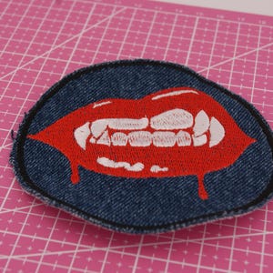 May include: Embroidered patch featuring a red mouth with fangs and dripping blood, set against a white tooth design. The patch is on a denim blue background with a black border. A unique accessory for clothing or bags.