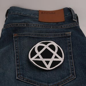 Può includere: Jeans blu scuro con una toppa ricamata in bianco e nero sulla tasca posteriore. La toppa presenta un pentagramma bianco all'interno di un cerchio nero, circondato da un design a forma di cuore bianco. Un'etichetta in pelle marrone con "& DENIM" è visibile.