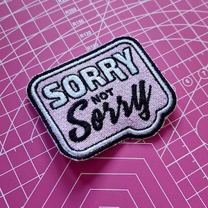May include: An embroidered patch with the text "SORRY NOT SORRY". The patch has a light pink background, black and white lettering, and a black border. It is set on a pink cutting mat.
