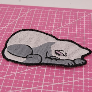 May include: Embroidered patch of a sleeping cat in grey and white, with a black outline. The cat is depicted lying down with closed eyes. The patch is on a pink cutting mat.