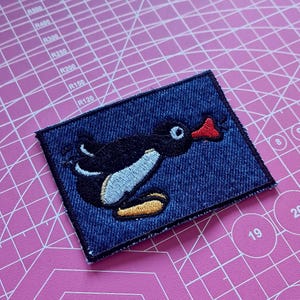 Può includere: Toppa in denim con un pinguino dei cartoni animati ricamato. Il pinguino ha un corpo nero, pancia bianca, becco rosso e piedi gialli. La toppa è rettangolare con un bordo nero.