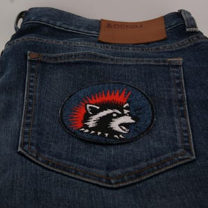 May include: Dark blue denim jeans with a raccoon patch on the back pocket. The patch features a black and white raccoon with a red mohawk on a blue oval background. A brown leather patch with the text "& DENIM" is visible.