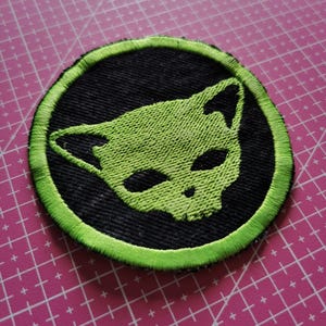 May include: Circular embroidered patch with a green cat face design on a black base. The cat's face has large eyes and pointed ears. The patch is edged with a bright green border. Suitable for application on clothing or accessories.