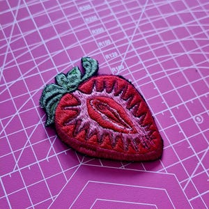 Könnte beinhalten: Gestickter Aufnäher in Form einer halben Erdbeere. Der Aufnäher hat ein detailliertes Design mit roten und rosa Nähten für die Frucht und grünen für die Blätter. Der Aufnäher ist wahrscheinlich für Kleidung oder Accessoires.