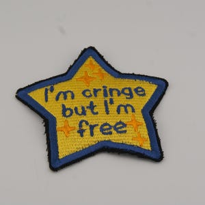May include: A yellow star-shaped embroidered patch with a blue border. The text "I'm cringe but I'm free" is in blue. Orange star accents and a black stitched edge complete the design.