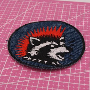 Könnte beinhalten: Runder, gestickter Aufnäher mit einem Waschbären mit rotem Irokesenschnitt und grimmigem Gesichtsausdruck. Der Aufnäher hat einen blauen Jeans-Hintergrund und schwarz-weiße Details. Ein Accessoire im Punkrock-Stil.