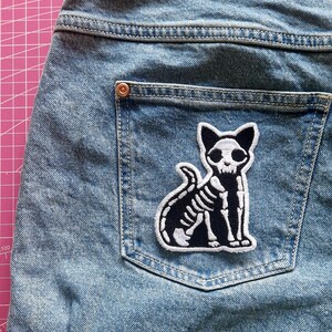 May include: Light blue denim jeans with a back pocket featuring a black and white embroidered patch. The patch depicts a skeleton cat with a detailed rib cage and skull. The jeans have a classic five-pocket design.