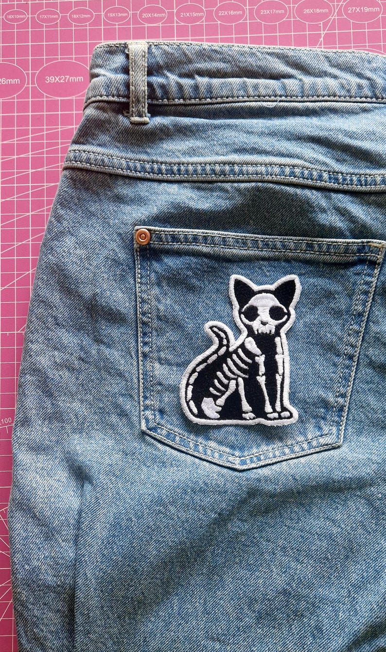 May include: Light blue denim jeans with a back pocket featuring a black and white embroidered patch. The patch depicts a skeleton cat with a detailed rib cage and skull. The jeans have a classic five-pocket design.