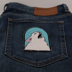 Op de afbeelding: Donkerblauwe denim jeans met een patch met een witte kat met open mond tegen een lichtblauwe achtergrond. De jeans heeft een bruine leren patch met de tekst "& DENIM".