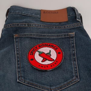 May include: Close-up of blue denim jeans with a brown leather patch and a circular patch on the back pocket. The patch features a red chili pepper design with the words "Spicy Romance" and "Snuy Bluv".