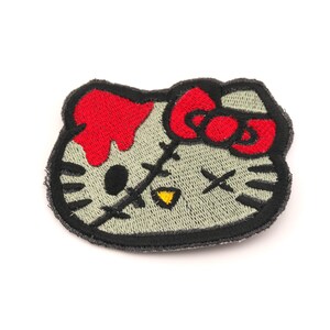 May include: Embroidered patch featuring a zombie-themed cartoon cat with a red bow and blood dripping from its head. The cat's eyes are crossed out, and it has a stitched mouth. The patch has a black border and a light green face.