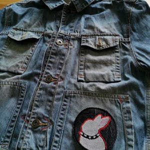 May include: Light blue denim jacket with multiple pockets and button closures. Features a round patch with a white dog silhouette wearing a studded collar. Red stitching accents the pockets and seams.