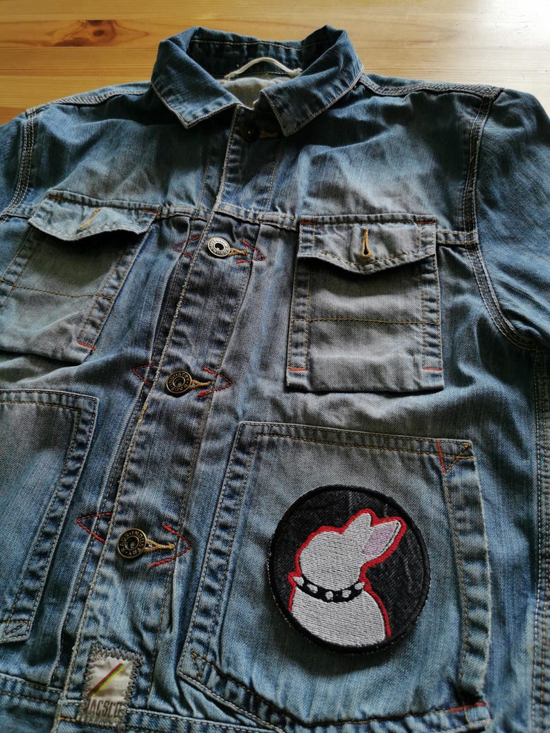 May include: Light blue denim jacket with multiple pockets and button closures. Features a round patch with a white dog silhouette wearing a studded collar. Red stitching accents the pockets and seams.