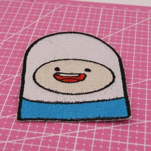Peut inclure: Écusson brodé représentant Finn de Adventure Time. Le patch a un haut blanc, un bas bleu clair et un visage beige avec une bouche souriante. Le patch est bordé de noir et est sur un tapis de coupe rose.