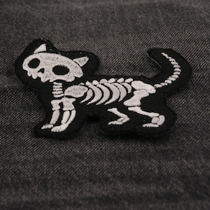Könnte beinhalten: Ein schwarzer, katzenförmiger Aufnäher mit einem weißen Skelett-Design. Der Aufnäher zeigt eine detaillierte Darstellung des Skeletts einer Katze, einschließlich Schädel, Rippen und Wirbelsäule. Der Aufnäher ist wahrscheinlich aus Stoff und zum Aufnähen oder Aufbügeln auf Kleidung oder Accessoires bestimmt.