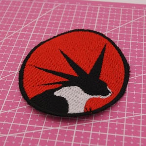 Peut inclure: Écusson brodé avec un motif de créature noir et blanc sur fond rouge. Le patch a une bordure noire et est posé sur un tapis de coupe rose.
