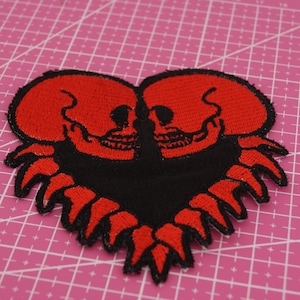 Può includere: Toppa ricamata con due teschi rossi che formano un cuore. Il cuore è delineato in nero con dettagli rossi a forma di fiamma. La toppa è su uno sfondo rosa a griglia.
