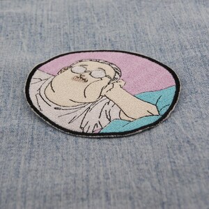 May include: Embroidered patch with a cartoon character wearing glasses, resting their chin on their hand. The patch has a black border, with a pink and blue background. The character is in a white robe.