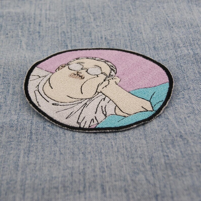 May include: Embroidered patch with a cartoon character wearing glasses, resting their chin on their hand. The patch has a black border, with a pink and blue background. The character is in a white robe.