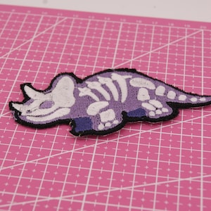 May include: An embroidered Triceratops dinosaur patch in purple and white, with a black border. The design features a skeleton, set against a pink grid. Ideal for customising clothing and accessories.