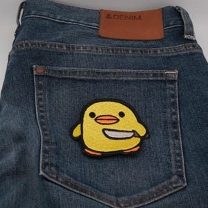 以下が含まれることがあります： バックポケットに黄色のヒヨコパッチが付いた青いデニムジーンズのクローズアップ。パッチにはナイフを持った漫画のヒヨコが描かれています。ジーンズには「& DENIM」と書かれた茶色の革ラベルが付いています。
