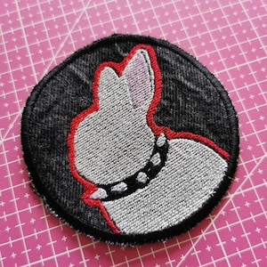 Pode incluir: Patch bordado com um design de coelho branco com contorno vermelho e uma coleira preta com tachas. O patch tem um fundo preto semelhante ao denim e uma borda preta. O patch tem formato circular.
