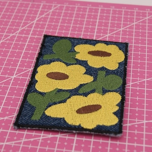 Può includere: Toppa ricamata con un motivo floreale di girasoli gialli, centri marroni e foglie verdi su sfondo denim. La toppa ha un bordo nero ed è di forma rettangolare. Probabilmente per abbigliamento o accessori.