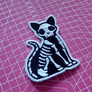 Könnte beinhalten: Schwarz-weißer, gestickter Aufnäher mit einem Katzen-Skelett-Design. Der Aufnäher zeigt eine Katze in sitzender Position mit eingerolltem Schwanz. Der Schädel der Katze ist sichtbar, und der Aufnäher hat einen weißen Rand.