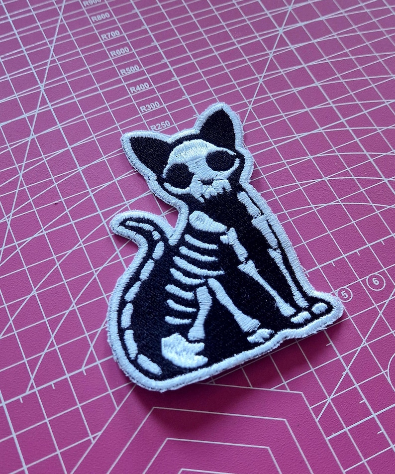 May include: Black and white embroidered patch featuring a cat skeleton design. The patch shows the cat in a sitting position with its tail curled. The cat's skull is visible, and the patch has a white border.