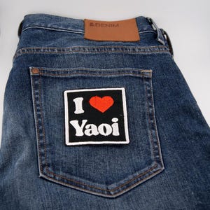 May include: Blue denim jeans with a patch on the back pocket. The patch is black with the words "I <3 Yaoi" in white and a red heart. The jeans have a brown leather tag that says "&DENIM".