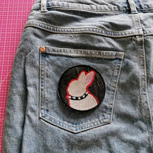 May include: Light blue denim jeans with a back pocket featuring a round patch. The patch has a white rabbit design with a red outline and a studded collar on a black background.