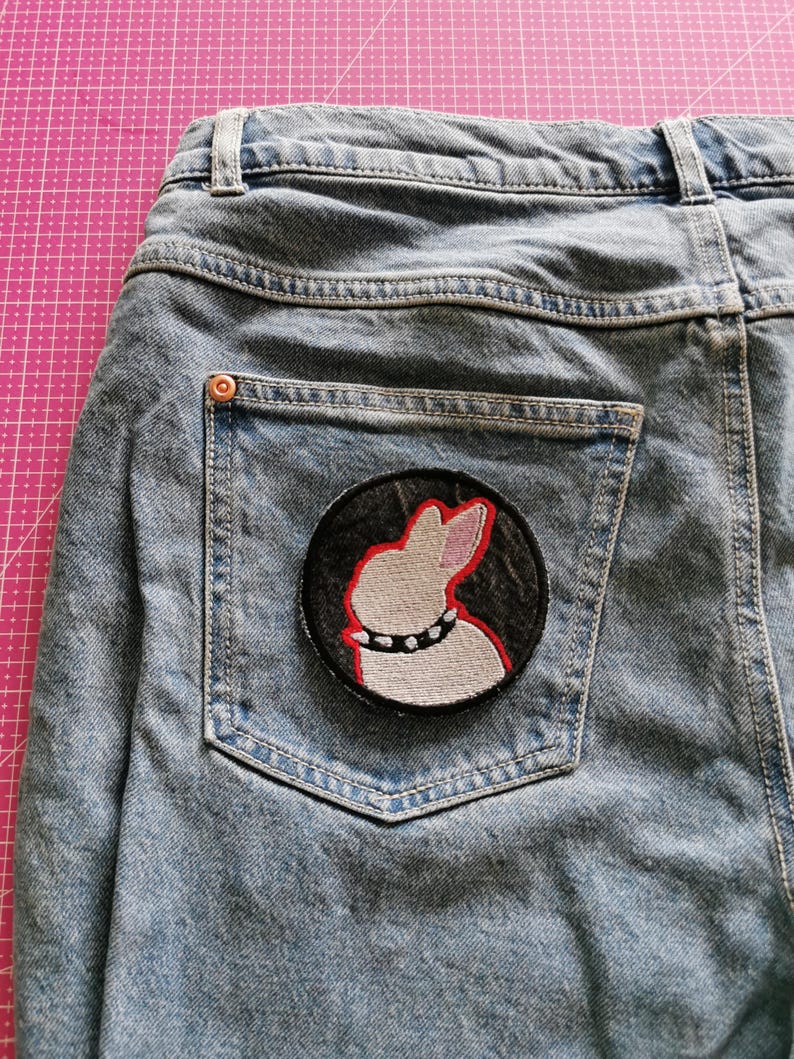 May include: Light blue denim jeans with a back pocket featuring a round patch. The patch has a white rabbit design with a red outline and a studded collar on a black background.