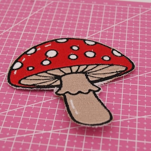 Può includere: Toppa ricamata a forma di fungo, con cappello rosso a pois bianchi e gambo beige. Il contorno è nero. La toppa è posta su uno sfondo rosa a griglia. Ideale per personalizzare abbigliamento e accessori.