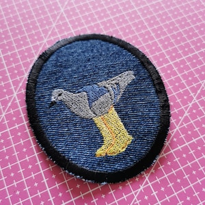 Può includere: Toppa ricamata rotonda con un disegno di uccello. La toppa ha uno sfondo blu denim con un bordo nero. L'uccello è raffigurato in grigio e blu, con stivali gialli. Adatta per abbigliamento.