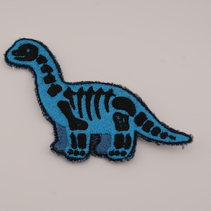 Könnte beinhalten: Ein blaues, gesticktes Abzeichen mit einem Dinosaurierskelett-Design. Der Dinosaurier ist hellblau mit schwarzen Details, einschließlich Brustkorb, Wirbelsäule und Kopf. Das Abzeichen hat ein strukturiertes Aussehen und einen dunkelblauen Rand.
