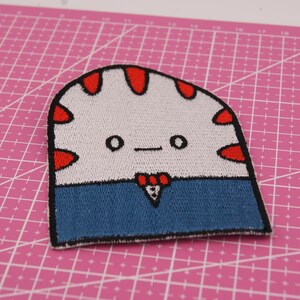 May include: Embroidered patch of a cartoon character with a white head, red details, and a blue body. The character has a simple face with two eyes and a small bow tie. The patch is on a pink cutting mat.