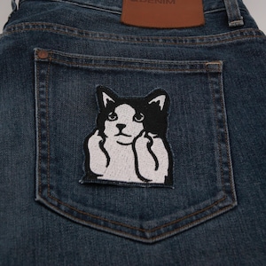May include: Dark wash denim jeans with a black and white cat patch on the back pocket. The patch features a cat with its paws up. A brown leather patch is visible at the waistband.