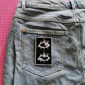 May include: Light blue denim jeans with a black patch featuring two stylized white eye designs. The patch is rectangular and sewn onto the back pocket of the jeans. The jeans have a classic five-pocket design.