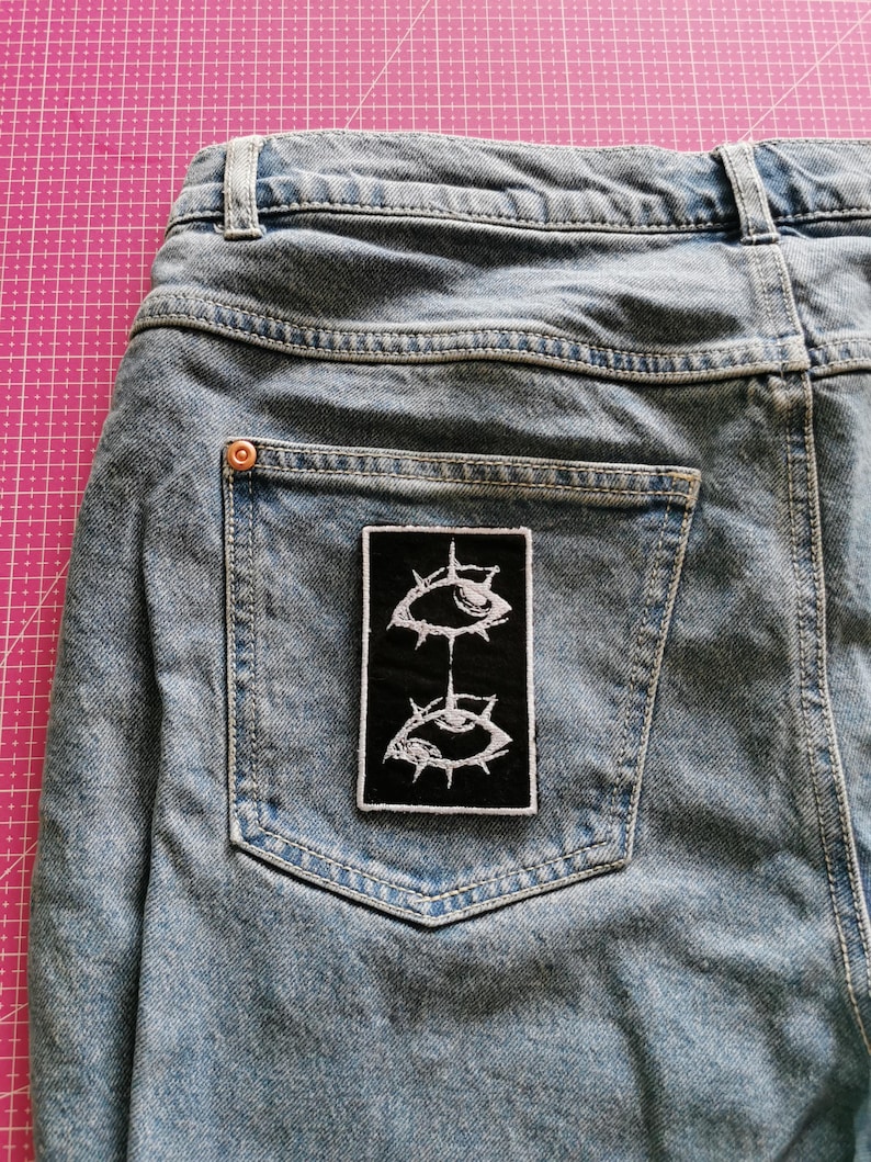 May include: Light blue denim jeans with a black patch featuring two stylized white eye designs. The patch is rectangular and sewn onto the back pocket of the jeans. The jeans have a classic five-pocket design.